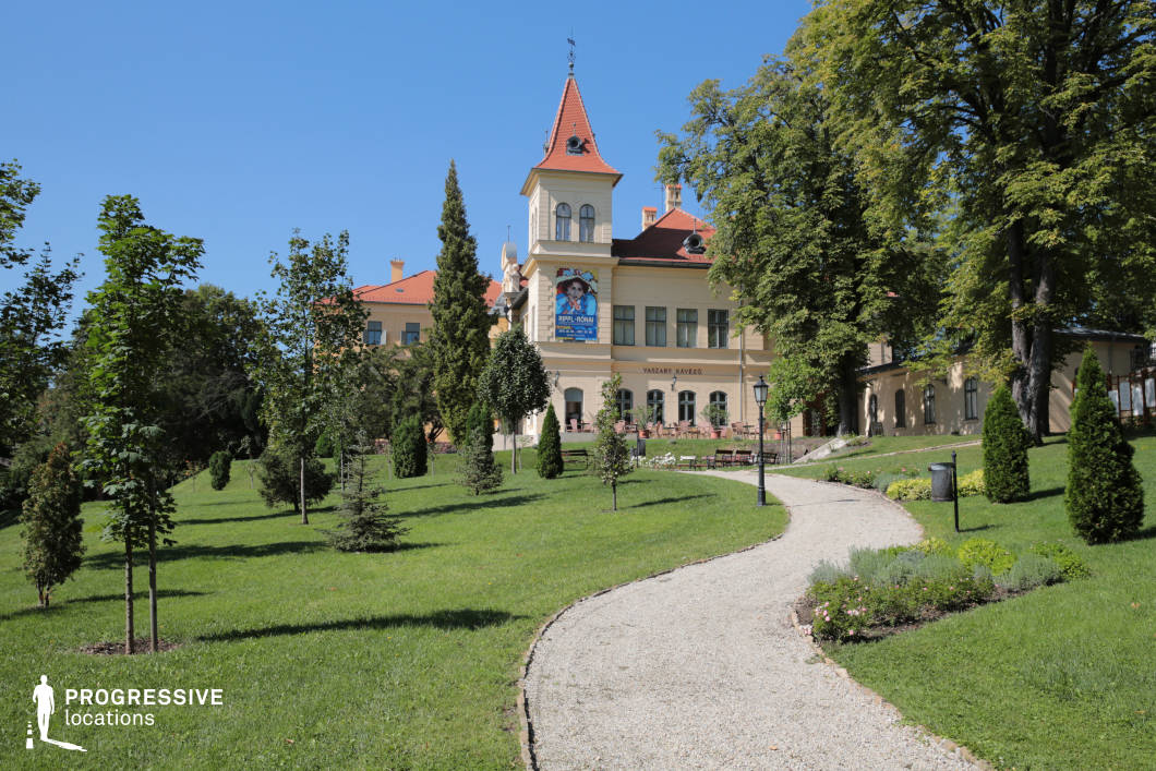 This image features a charming historic villa with warm-toned walls, red-tiled roofs, and a prominent tower, nestled within a lush landscaped garden. A gently winding gravel pathway leads towards the building, framed by manicured greenery and vintage-style lampposts, enhancing the inviting and picturesque ambiance. The villa’s classic European architecture, with arched windows and decorative details, blends harmoniously with the surrounding natural elements. This location is ideal for filming period dramas, romantic films, or lighthearted character-driven narratives, offering a tranquil and visually refined setting.