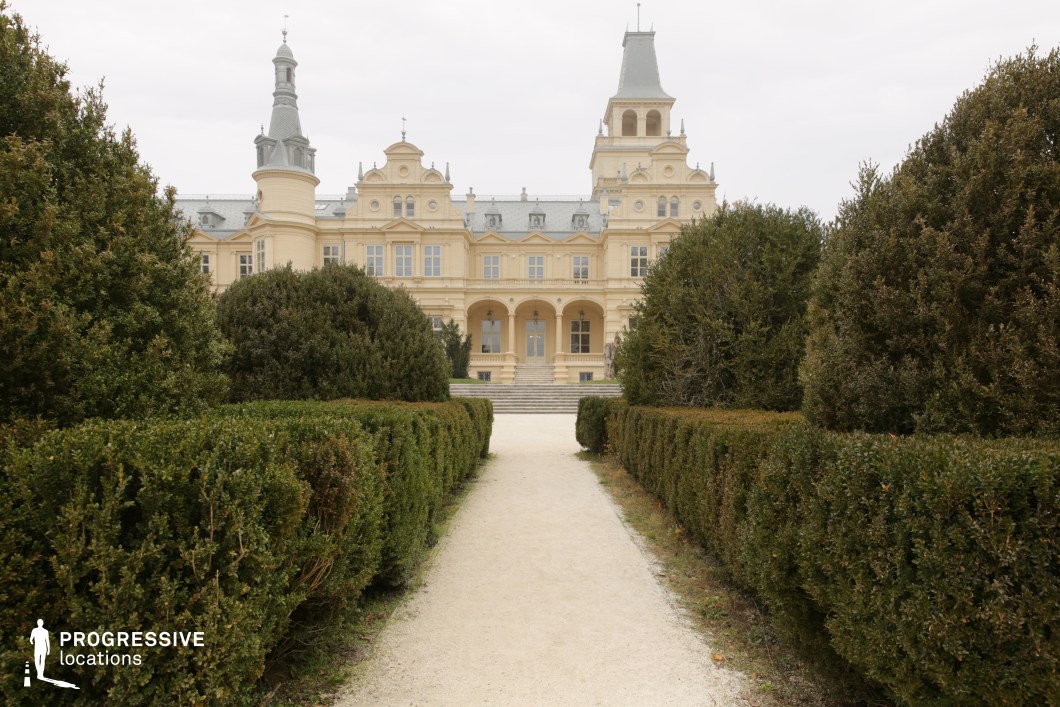 This image features an opulent historic palace framed by a meticulously maintained garden, with a symmetrical pathway leading toward the grand entrance. The architectural design blends neoclassical and baroque elements, with ornate facades, arched balconies, and towering spires that add a regal presence. The soft yellow exterior, adorned with sculpted reliefs and decorative moldings, enhances the building's stately elegance. The structured hedges and tree-lined approach reinforce the palace’s aristocratic charm. This location is ideal for filming period dramas, historical romances, or royal narratives, providing a sophisticated and picturesque setting for scenes of nobility, diplomacy, or intrigue.