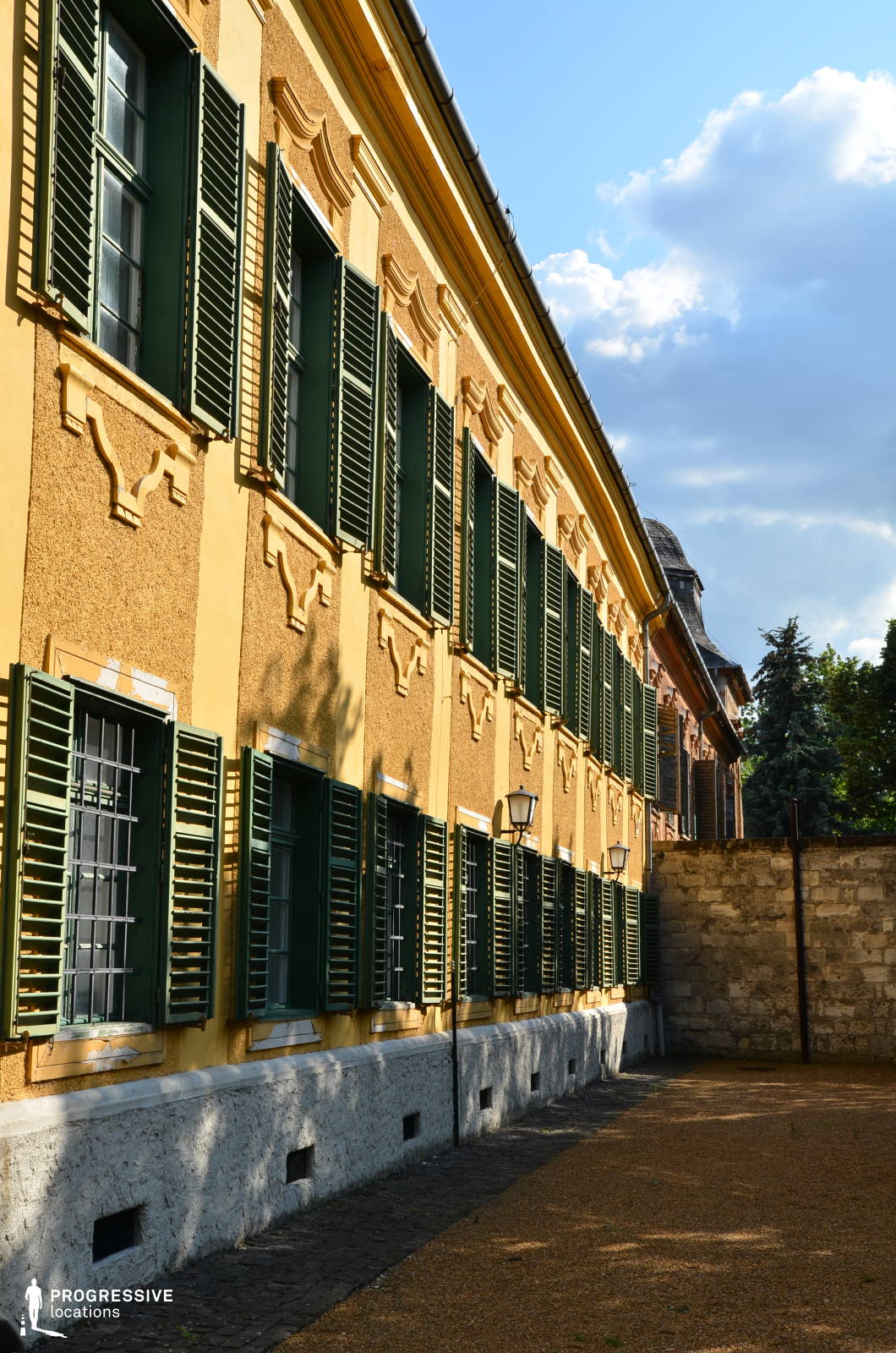 This elegant historic building features a warm yellow facade with decorative stucco elements and dark green wooden shutters, creating a striking contrast. The repetitive pattern of windows, framed with ornamental details, enhances the classical symmetry of the structure. The shutters, some fully open and others partially closed, add depth and texture to the exterior. The lower portion of the building is accentuated by a white stone foundation, while a narrow courtyard paved with gravel and brick runs along the side, enclosed by a rustic stone wall. This location is ideal for period films, historical dramas, and European-set productions, providing a visually rich and atmospheric backdrop.