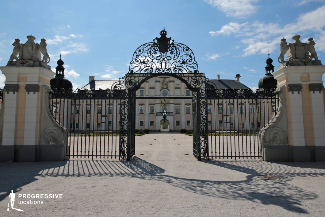 This image showcases a grand Baroque palace framed by an elaborate wrought-iron gate with intricate scrollwork and heraldic emblems. The symmetrical façade of the palace features soft pastel hues, decorative pilasters, and a central pediment, embodying classical elegance. The imposing entrance leads to a vast open courtyard, emphasizing formality and prestige. The structured layout, combined with the play of light and shadow on the gravel path, enhances the depth and regal ambiance of the setting. This location is ideal for filming historical dramas, period pieces, or aristocratic-themed productions, offering a sophisticated and visually striking backdrop.