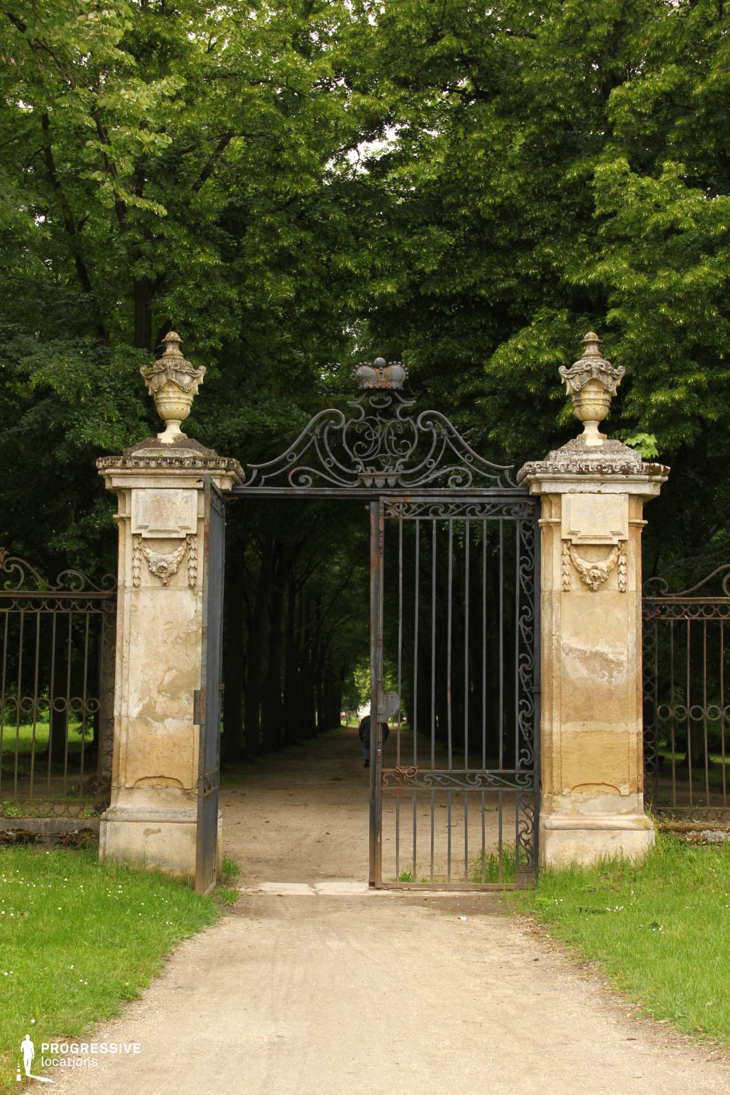 This grand wrought-iron gate, framed by weathered stone pillars, serves as an elegant entrance to a tree-lined pathway. The intricate ironwork, adorned with delicate scroll patterns and a regal crest, adds a sense of refinement and historical significance. Stone urns rest atop the gateposts, contributing to the classical aesthetic. Beyond the partially open gate, a straight dirt path disappears into a dense canopy of trees, creating a sense of mystery and seclusion. This location is ideal for historical dramas, period films, and gothic or mystery-themed productions, offering a visually compelling and atmospheric setting.