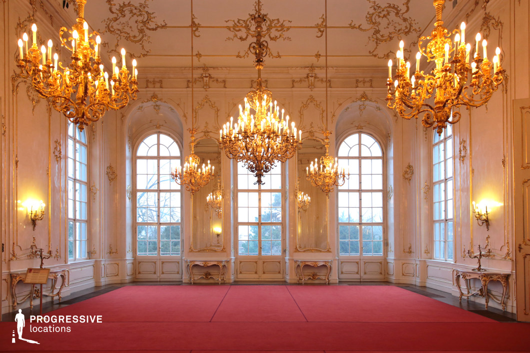 This stunning Baroque-style ballroom exudes regal elegance, featuring high arched windows that flood the space with natural light, accentuating the intricate gilded wall moldings and delicate stucco decorations. The symmetrical arrangement of grand crystal chandeliers casts a warm glow over the room, enhancing the lavish atmosphere. A red carpet stretches across the polished floor, adding a sense of formality and grandeur. Large mirrors framed in gold detailing further amplify the opulence, creating a refined and sophisticated setting. This location is ideal for filming historical dramas, royal court scenes, high-society gatherings, and period-piece productions that require an aristocratic and luxurious ambiance.