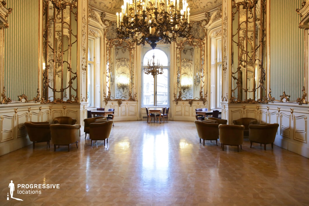 This stunning Rococo-style hall radiates elegance with its gilded moldings, large arched windows, and tall mirrors that create a sense of spaciousness and grandeur. The pastel green and gold-accented walls, combined with intricate carvings and floral motifs, add a refined charm, while the polished parquet floor reflects the warm glow of the elaborate chandeliers. The arrangement of plush velvet armchairs and dark wooden tables suggests a luxurious social setting, ideal for aristocratic gatherings or formal events. This location is perfect for filming high-society period dramas, classical romance, or regal intrigue in genres such as historical fiction, costume dramas, and sophisticated thrillers.