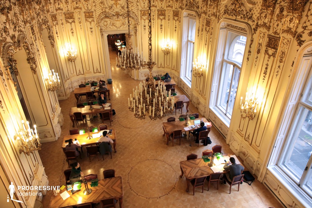 This opulent Rococo-style hall blends grandeur with intellectual refinement, featuring intricate gilded moldings, soaring windows, and an array of chandeliers that cast a warm glow over the parquet flooring. The high, elaborately decorated ceiling enhances the sense of space, while the symmetrical arrangement of wooden tables and green-shaded lamps creates an atmosphere of scholarly elegance. Large mirrors reflect the delicate detailing of the walls, adding depth and luminosity to the room. This location is ideal for filming aristocratic salons, intellectual debates, or period dramas set in academia, making it a perfect backdrop for historical fiction, classic literary adaptations, or refined mystery narratives.