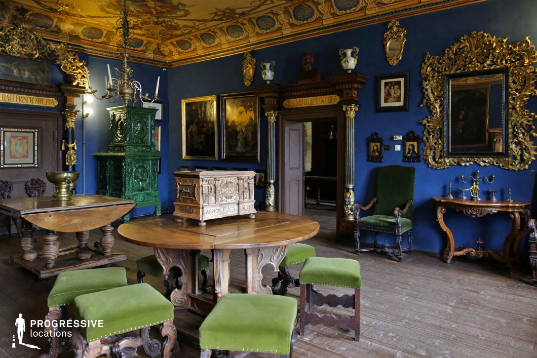 This strikingly opulent room blends Renaissance and Baroque influences, featuring deep blue walls adorned with gilded mirrors, elaborate picture frames, and classical artwork. The richly carved wooden furniture, including tables with intricate detailing and green velvet-upholstered stools, adds an aristocratic character. A grand green ceramic stove stands as a focal point, enhancing the historical ambiance. The ceiling, decorated with gold leaf motifs and heraldic elements, elevates the space’s grandeur. This location is ideal for filming period dramas, historical mysteries, or fantasy productions requiring an atmosphere of nobility, secrecy, or old-world intrigue.