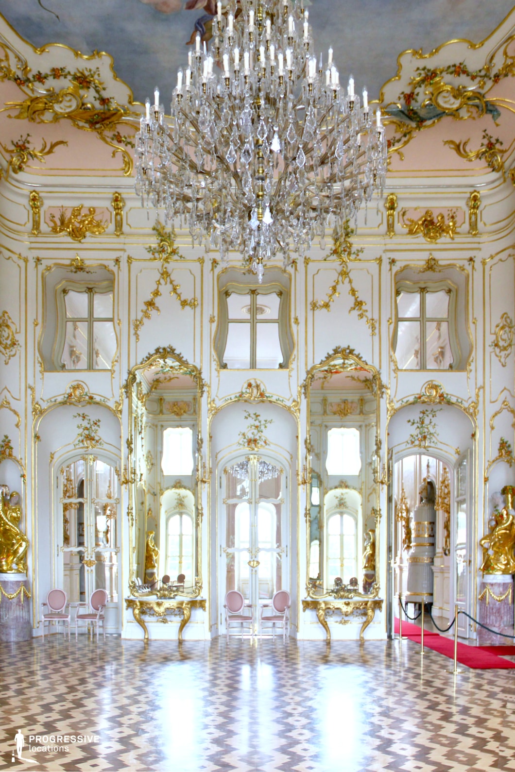 This stunning Rococo-style ballroom is adorned with elaborate gold moldings, mirrored wall panels, and intricate floral motifs, creating an atmosphere of grandeur and refinement. The vast, checkered parquet floor enhances the room's symmetry, leading the eye toward arched doorways framed by ornate gilded sculptures. A grand crystal chandelier hangs from a ceiling decorated with delicate frescoes, casting a soft, opulent glow throughout the space. Tall windows allow natural light to reflect off the mirrors, amplifying the sense of openness and luxury. This location is ideal for filming historical dramas, period romances, or regal settings, offering an exquisite and visually captivating backdrop for scenes of aristocratic elegance and lavish gatherings.