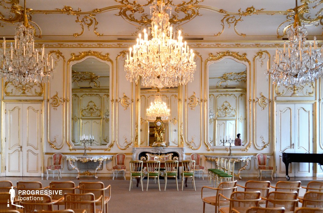This breathtaking Rococo ballroom showcases an opulent design with intricate gold moldings, large arched mirrors, and grand crystal chandeliers that amplify the sense of luxury. The white and gold palette creates an airy yet regal atmosphere, while the ornate ceiling detailing enhances the grandeur. A marble fireplace, delicate carved furniture, and a long ceremonial table suggest a space designed for aristocratic gatherings and high-society events. The mirrored reflections add depth, making the room appear even more expansive. This location is ideal for filming period dramas, royal ceremonies, or elegant social affairs in historical fiction, classic romance, or refined intrigue.