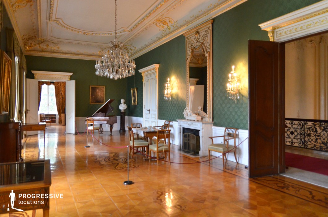 This elegant neoclassical salon blends aristocratic charm with a warm, intimate ambiance. Deep green damask-patterned walls contrast beautifully with intricate white and gold moldings, while a grand chandelier and ornate wall sconces illuminate the polished parquet flooring. A stately marble fireplace, complete with sculptural detailing and an oversized mirror, adds to the room’s grandeur. Period furniture, including a set of finely crafted chairs and a central table, contributes to the refined aesthetic. A grand piano in the background reinforces the salon’s historical significance as a space for music and cultural gatherings. This location is ideal for filming aristocratic salons, classical music performances, or period dramas, fitting for historical fiction, refined literary adaptations, or artistic intrigue.