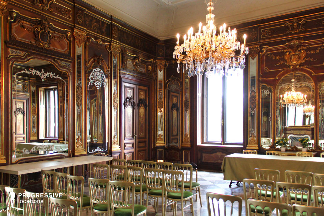 This extravagant Baroque-style hall is adorned with richly carved wooden panels, large gilded mirrors, and intricate gold leaf detailing that enhance the sense of opulence and grandeur. A crystal chandelier hangs from the high ceiling, casting a warm glow over the marble-tiled floor and the elegant green-upholstered chairs arranged in a formal setup. The interplay of mirrors creates an illusion of depth and infinite reflections, amplifying the luxurious atmosphere. Tall windows allow natural light to illuminate the finely decorated walls, while ornamental motifs and intricate engravings add to the room’s historical richness. This location is ideal for filming aristocratic gatherings, diplomatic events, or high-stakes confrontations in historical dramas, period pieces, or regal thrillers.