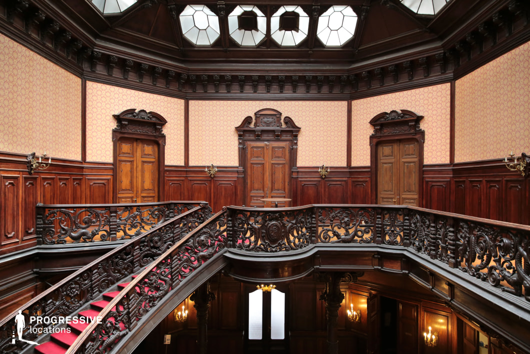 This lavish interior features a grand, curved staircase with an elaborately carved wrought-iron balustrade and a deep red carpet runner, adding a regal touch. The dark wood paneling extends across the walls, contrasted by vintage-patterned wallpaper, creating a warm yet imposing atmosphere. Above, an octagonal skylight allows natural light to filter into the space, highlighting the architectural details. Multiple wooden doors with decorative pediments line the upper level, enhancing the room’s symmetrical and formal composition. This location is perfect for filming period dramas, gothic thrillers, or aristocratic intrigue scenes, offering a visually rich and historically immersive setting.