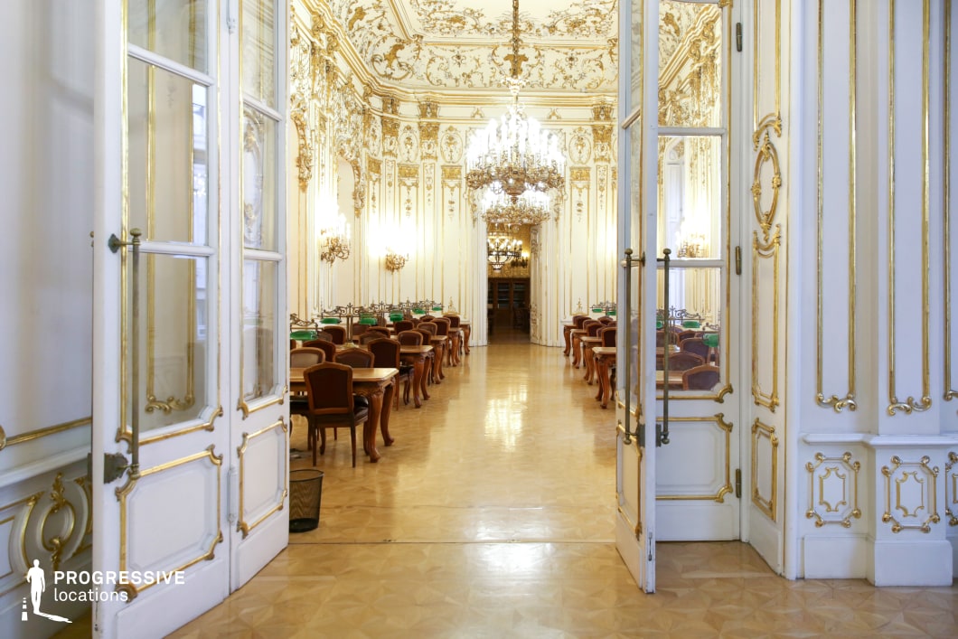 This stunning Rococo-style hall is a masterpiece of classical elegance, featuring soaring ceilings adorned with intricate gold moldings and delicate floral motifs. Grand chandeliers hang symmetrically, illuminating the white and gold-paneled walls, which are further enriched by tall mirrors reflecting the opulent interior. The polished parquet flooring enhances the hall’s refined atmosphere, while the symmetrical arrangement of wooden tables and chairs, accented with green desk lamps, adds a scholarly yet aristocratic charm. The grand double doors open to an extended view of the lavish space, adding depth and continuity. This location is ideal for filming period dramas, high-society gatherings, or refined intellectual discussions in genres such as historical fiction, classic romance, or political intrigue.