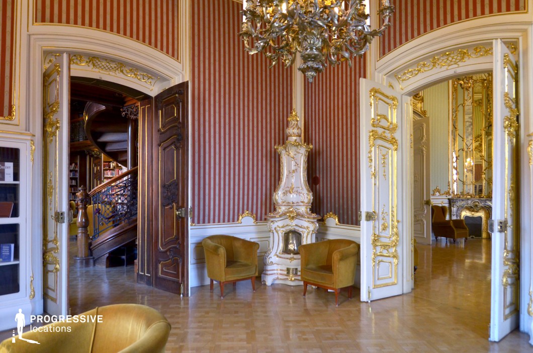 This exquisitely designed Rococo-style salon radiates aristocratic elegance, featuring red-striped wallpaper accented with gold detailing, intricate white and gold moldings, and a lavish ceramic fireplace as a central focal point. The grand double doors, framed with gilded carvings, open into a luxurious adjoining room with tall mirrors and an opulent fireplace, enhancing the depth and continuity of the space. To the left, a dark wooden doorway reveals a grand spiral staircase leading to an ornate library, adding an air of scholarly refinement. The plush mustard-colored armchairs contrast beautifully with the polished parquet flooring, creating a regal yet inviting atmosphere. This location is ideal for filming aristocratic gatherings, intellectual debates, or clandestine meetings in historical dramas, political thrillers, and classic literary adaptations.