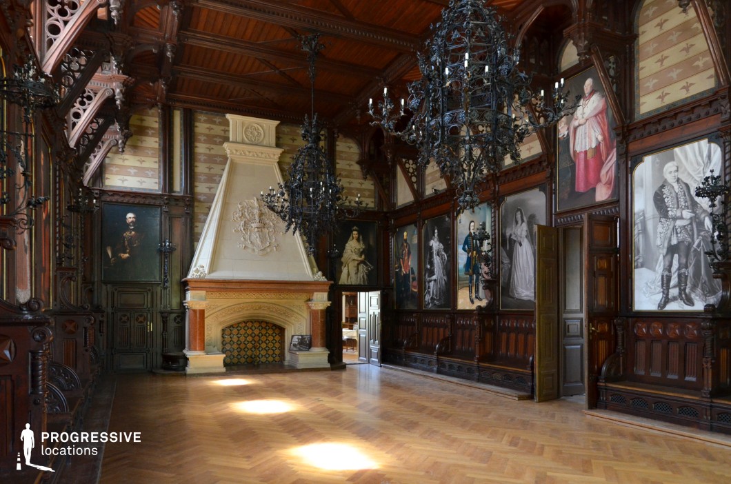 This stately hall, rich in Gothic Revival details, features dark wood paneling, a soaring coffered ceiling, and elaborately crafted wrought-iron chandeliers that enhance its imposing ambiance. The walls are adorned with large-scale historical portraits framed by intricate wood carvings, contributing to the room’s aristocratic grandeur. A monumental stone fireplace, crowned with an ornate coat of arms, serves as the focal point, with its tiled hearth adding a warm contrast to the otherwise dark-toned interior. The herringbone-patterned parquet floor reflects the soft natural light filtering through. This location is ideal for filming period dramas, historical epics, Gothic mysteries, and aristocratic intrigue, offering an atmosphere of prestige and dramatic intensity.