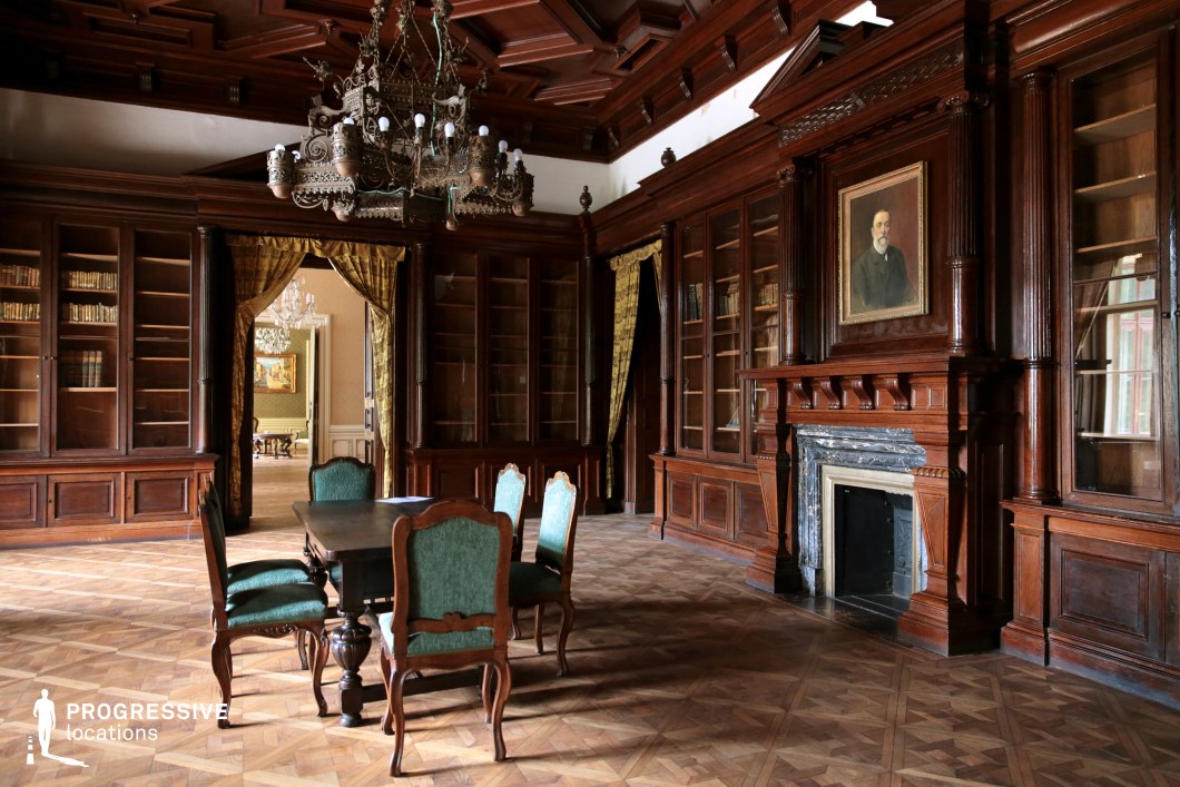This stately library exudes old-world elegance, featuring floor-to-ceiling wooden bookshelves, a grand fireplace with a marble mantel, and an intricately carved ceiling that enhances the room’s architectural richness. A large antique chandelier hangs at the center, casting a warm, moody glow over the parquet flooring and richly upholstered furniture. The deep wood tones, velvet drapery, and finely detailed moldings create an atmosphere of intellect and refinement. The adjoining room, visible through the open doorway, adds depth and continuity to the space. This location is ideal for filming period dramas, aristocratic settings, political thrillers, or mystery films requiring an academic, sophisticated, or secretive ambiance.