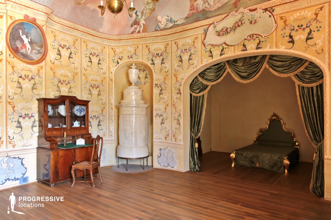 This elegant Rococo-style bedroom exudes historical charm, featuring delicate hand-painted walls adorned with floral and mythological motifs. The alcove bed, framed by rich green drapery with gold accents, adds a sense of intimacy and refinement, while the ornately gilded headboard enhances the aristocratic aesthetic. A large white ceramic stove stands as both a decorative and functional element, complementing the finely crafted wooden furnishings, including a writing desk with a display cabinet. The pastel ceiling fresco and warm wooden flooring create a harmonious, inviting atmosphere. This location is ideal for filming period dramas, historical romance, and aristocratic intrigue, offering an authentic and visually rich backdrop.