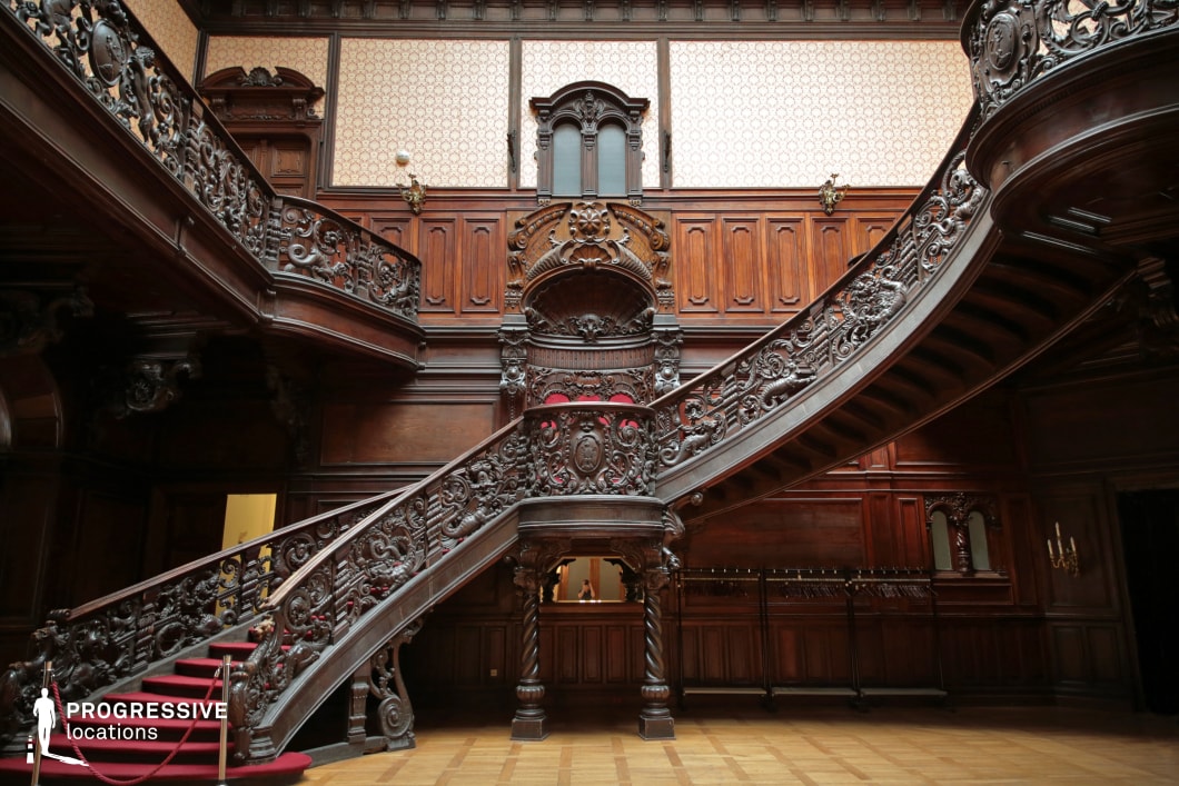 This opulent interior showcases a grand staircase with intricately carved dark wood balustrades, exuding a sense of luxury and historical depth. The polished wooden paneling extends across the walls, adorned with decorative moldings and subtle patterned wallpaper that enhances the classical ambiance. The sweeping dual staircase, lined with a deep red carpet, leads to an upper gallery with ornate railings, reinforcing the architectural grandeur. The dimly lit space, featuring antique-style sconces and an imposing wooden structure, creates a dramatic and refined atmosphere. This location is ideal for filming period dramas, aristocratic intrigue, Gothic-inspired narratives, or mystery thrillers that require an elegant yet imposing backdrop.