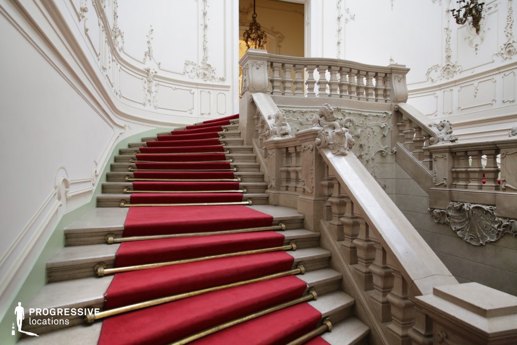 This majestic Baroque-style staircase exudes elegance with its sweeping curves, intricately carved balustrades, and symmetrical design. The stately white walls are adorned with delicate stucco detailing, enhancing the grandeur of the space. A plush red carpet with gold accents runs along the stone steps, reinforcing the aristocratic ambiance. Ornate wall-mounted chandeliers and an elaborate ceiling lantern add to the opulence, casting a soft glow over the architectural details. This location is ideal for filming dramatic entrances, high-society gatherings, or power struggles in historical dramas, period romances, or regal intrigue-filled thrillers.