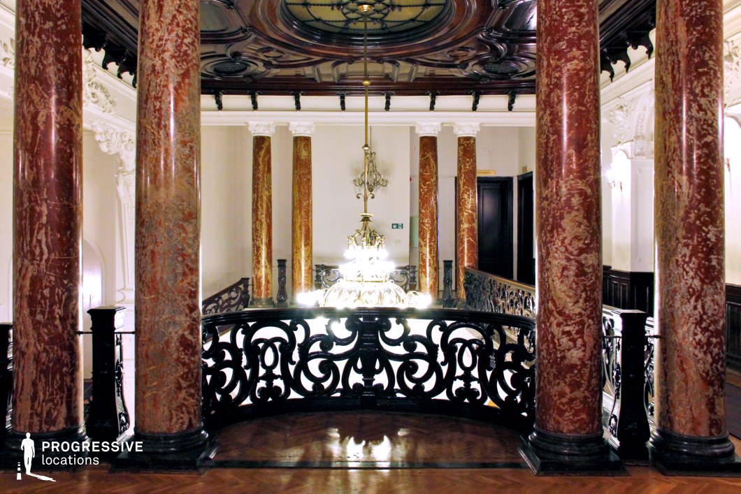 This luxurious gallery features striking red marble columns with black bases, supporting a richly detailed coffered ceiling with dark wood inlays. The ornate, wrought-iron balustrade frames a grand chandelier that serves as the focal point of the space, casting an elegant glow. The polished parquet flooring enhances the sense of refinement, while the cream-colored walls with intricate moldings contrast beautifully with the darker architectural elements. The symmetrical arrangement of the columns and the decorative interplay between materials create a regal and sophisticated atmosphere. This location is ideal for filming period dramas, high-society intrigue, or historical thrillers, offering a visually captivating and aristocratic setting.