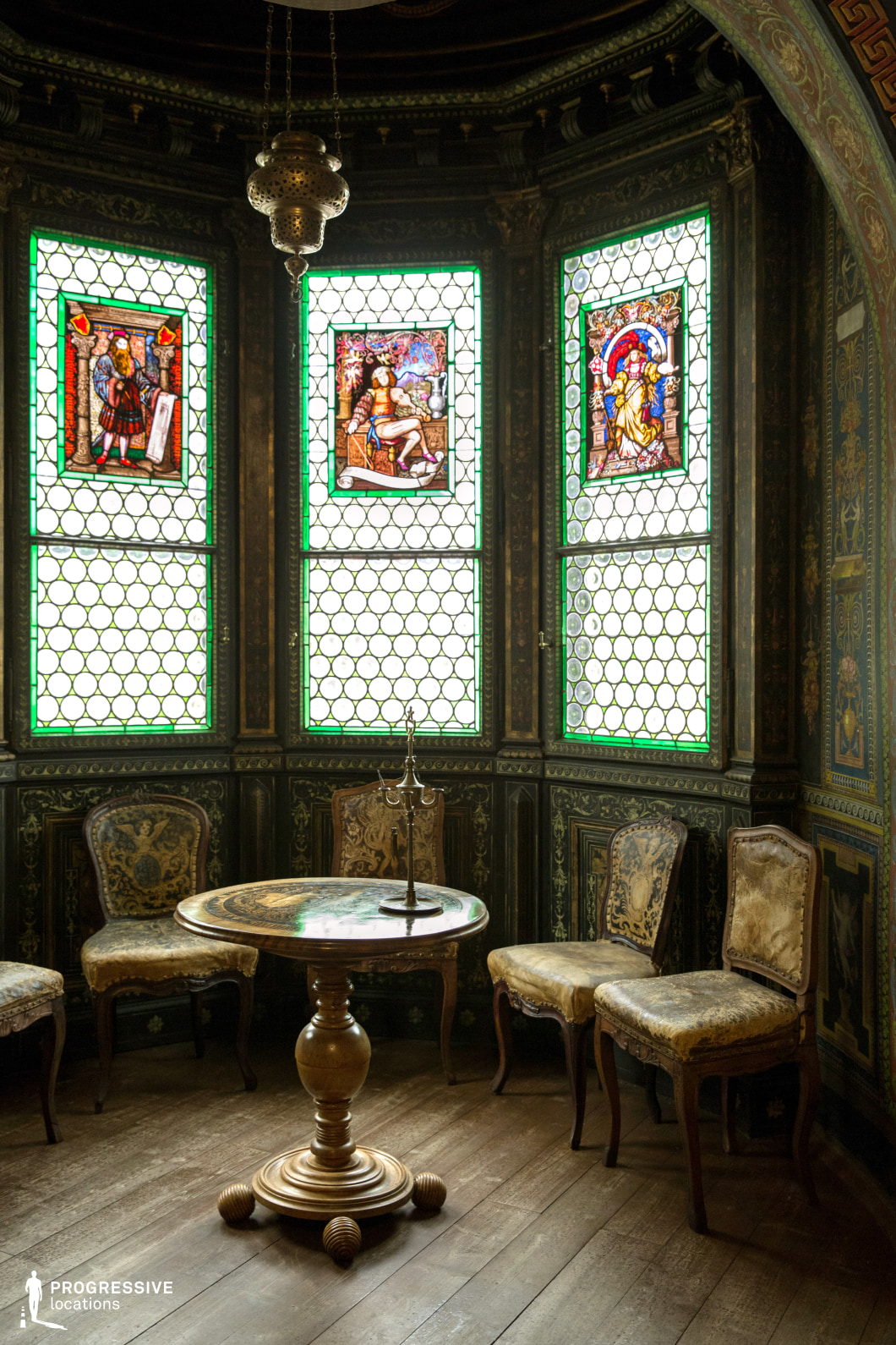 This intimate alcove exudes old-world opulence, featuring richly carved dark wood paneling, antique furnishings, and vibrant stained-glass windows depicting historical and mythological figures. The intricate geometric patterns and delicate floral motifs on the walls enhance the space’s Gothic Revival elegance. A small round table with an ornate pedestal base and aged velvet-upholstered chairs contribute to the room’s vintage charm. The dim lighting, complemented by the glow filtering through the colorful glass, creates a moody, atmospheric setting. This location is ideal for filming period dramas, historical intrigue, aristocratic settings, or scenes requiring a secretive, mystical, or scholarly ambiance.