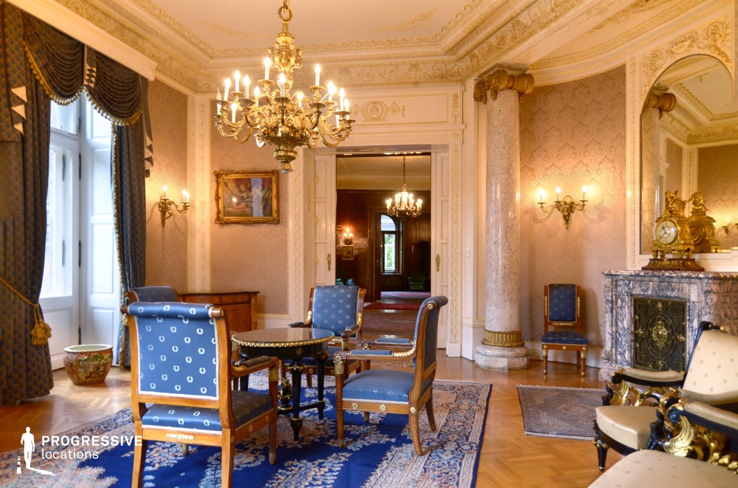 This refined neoclassical salon exudes sophistication with its richly decorated walls, intricate ceiling moldings, and a grand chandelier that casts a warm glow over the space. The room features elegant blue-upholstered furniture with gold detailing, a polished black marble fireplace adorned with a gilded clock, and a plush Persian-style rug that enhances the aristocratic ambiance. Towering marble columns and large mirrors framed in ornate gold add depth and grandeur, while the deep blue drapery provides a regal contrast to the soft-toned walls. The open doorway leads into another richly furnished room, creating a seamless flow between lavish interiors. This location is ideal for filming historical dramas, diplomatic meetings, or aristocratic intrigues, making it perfect for period pieces, refined thrillers, and classical literary adaptations.