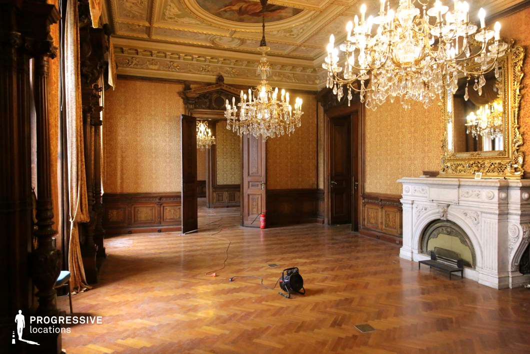 This opulent ballroom features intricate Baroque-style detailing, highlighted by gilded chandeliers, a marble fireplace with elaborate carvings, and dark wooden paneling that adds depth and warmth to the space. The richly patterned wallpaper complements the coffered ceiling, which showcases ornate stucco work and painted motifs. Large mirrors with gilded frames enhance the room’s grandeur, reflecting the light from the chandeliers. The parquet flooring, laid in a herringbone pattern, contributes to the historical elegance, while tall windows framed by heavy drapes allow natural light to filter through. This location is ideal for filming period dramas, aristocratic social gatherings, or mysterious, suspenseful scenes in genres such as historical fiction, gothic horror, or psychological thrillers.