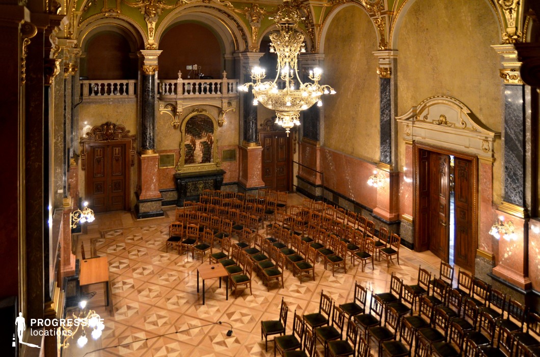 This stately Baroque-style hall features towering arched ceilings, rich marble columns, and intricate gold detailing, creating an atmosphere of historic grandeur. The parquet flooring, arranged in a geometric pattern, adds refinement to the space, while an imposing chandelier casts a warm glow, highlighting the opulent architectural details. The large wooden doors, adorned with intricate carvings, enhance the hall's sense of prestige. A decorative balcony with classical balustrades overlooks the main floor, adding depth to the composition. This location is well-suited for filming political or aristocratic gatherings, courtroom dramas, period pieces, or mystery thrillers requiring a regal and imposing setting.
