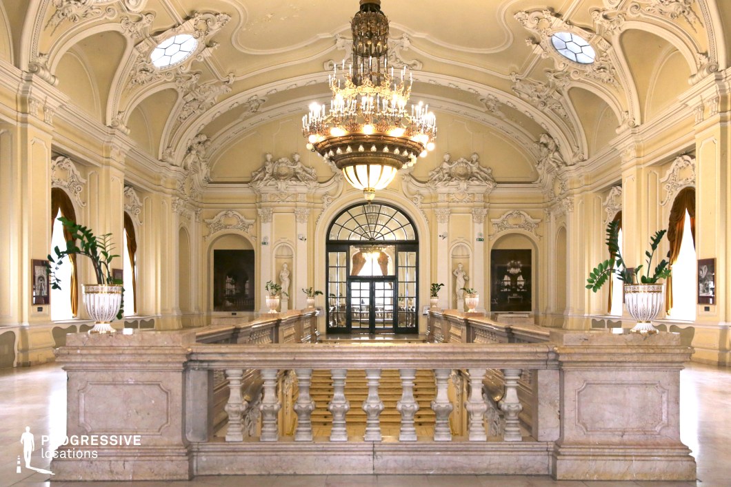 This opulent hall showcases intricate Baroque and Rococo architectural details, featuring a sweeping marble staircase, sculpted balustrades, and an elaborately decorated vaulted ceiling adorned with stucco reliefs. A grand chandelier dominates the space, casting warm light across the pale cream-colored walls, which are punctuated by arched windows draped with elegant curtains. Ornamental sculptures and classical urns enhance the refined aesthetic, while the symmetrical layout and arched glass doors create a sense of depth and grandeur. This location is ideal for filming historical dramas, period-piece romances, high-society gatherings, or regal courtly scenes that require an atmosphere of luxury and sophistication.