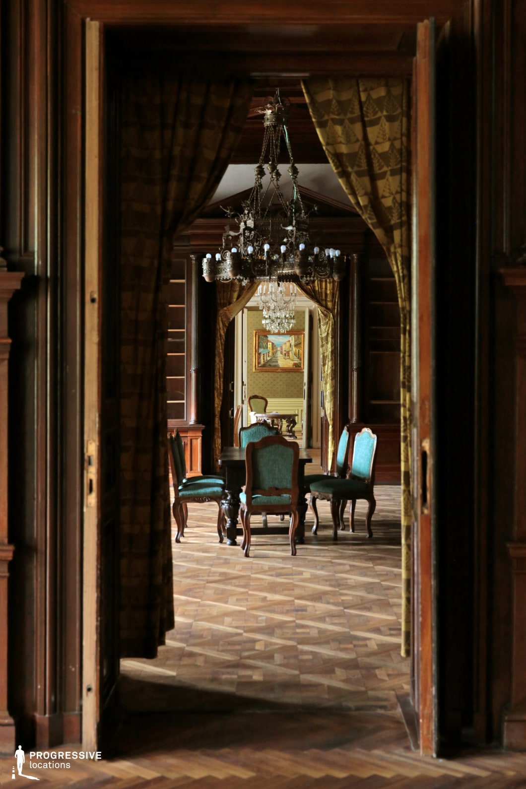 This grand salon features richly paneled wooden walls, an intricate parquet floor, and a series of chandeliers that create a dramatic sense of depth as they recede into multiple adjoining rooms. The dark wood contrasts beautifully with the deep green upholstered chairs, while heavy drapery frames the entrances, adding to the regal ambiance. The interplay of open doorways and reflections enhances the visual complexity of the space. This location is ideal for filming period dramas, aristocratic intrigue, historical mysteries, or intellectual debates in settings that require an atmosphere of refinement and sophistication.