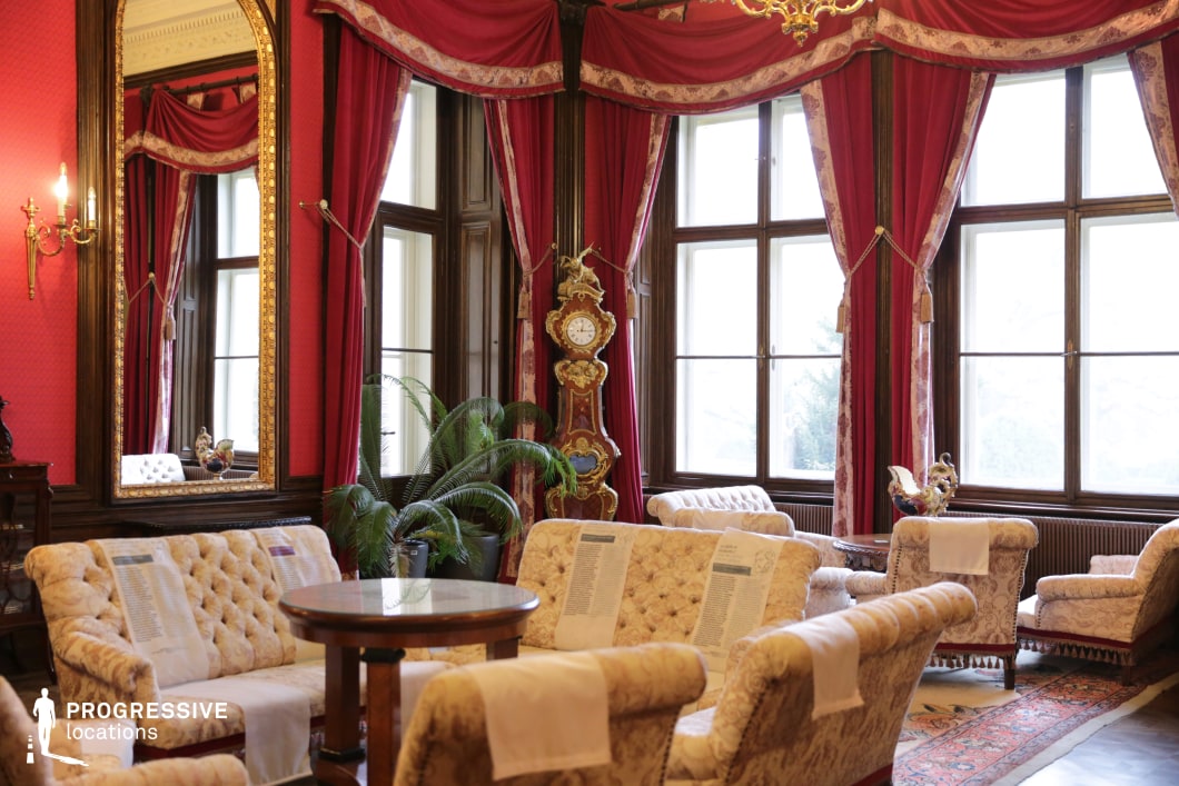 This opulent Victorian-style lounge exudes warmth and sophistication with its deep red damask wallpaper, heavy velvet drapery, and dark wood paneling. The room is furnished with plush, cream-colored tufted seating that contrasts elegantly with the dramatic backdrop. A gilded floor clock with intricate carvings serves as a centerpiece, reinforcing the historical grandeur. Large mirrors framed in gold reflect the chandeliers’ soft light, enhancing the room's richness. The expansive windows framed by ornate curtains add depth and natural light. This location is ideal for filming aristocratic gatherings, secretive discussions, or lavish period settings in historical dramas, literary adaptations, or political intrigue films.