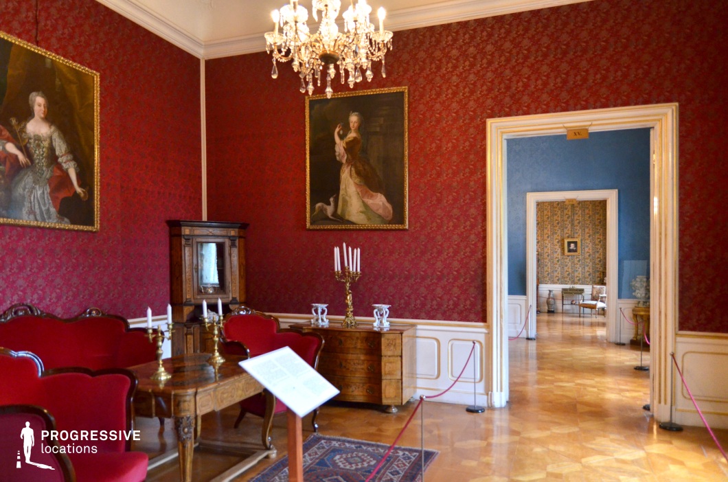 This lavish salon exudes royal elegance with its deep red damask wallpaper, gilded chandeliers, and antique furnishings. The richly upholstered seating in matching red tones creates a cohesive, opulent aesthetic, while the polished parquet flooring enhances the grandeur of the space. Large, gold-framed portraits depicting aristocratic figures contribute to the historical character of the room. A series of interconnected rooms with varying wall colors, visible through the ornate doorway, adds depth and visual contrast. This location is ideal for filming high-society gatherings, political intrigues, or period dramas, making it a perfect setting for historical fiction, regal thrillers, and classical literary adaptations.