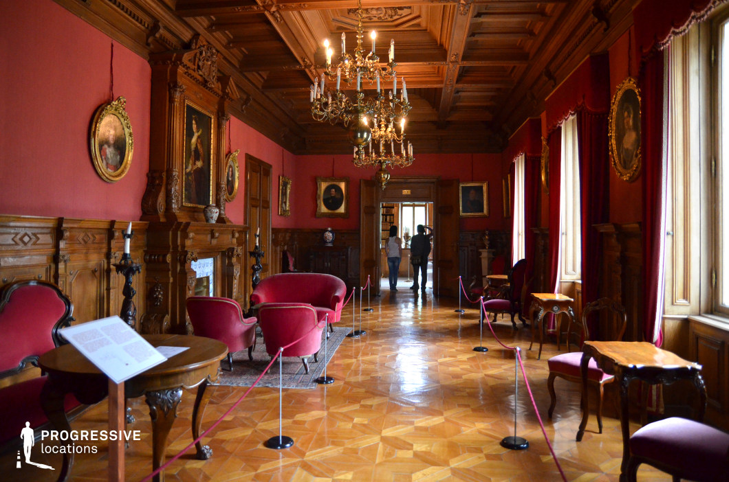 This opulent study room features deep red fabric-covered walls, richly carved wooden paneling, and an intricately coffered ceiling, creating an atmosphere of grandeur and historical refinement. Ornate gold-framed portraits line the walls, complementing the antique-style furniture, including red velvet armchairs and dark wooden tables. Large windows with heavy draped curtains allow soft natural light to accentuate the polished parquet flooring. Two elaborate chandeliers hang from the ceiling, enhancing the room’s warm glow. This location is ideal for filming period dramas, historical thrillers, or aristocratic intrigue, offering a regal and sophisticated setting for scenes of power, wealth, and intellectual discourse.