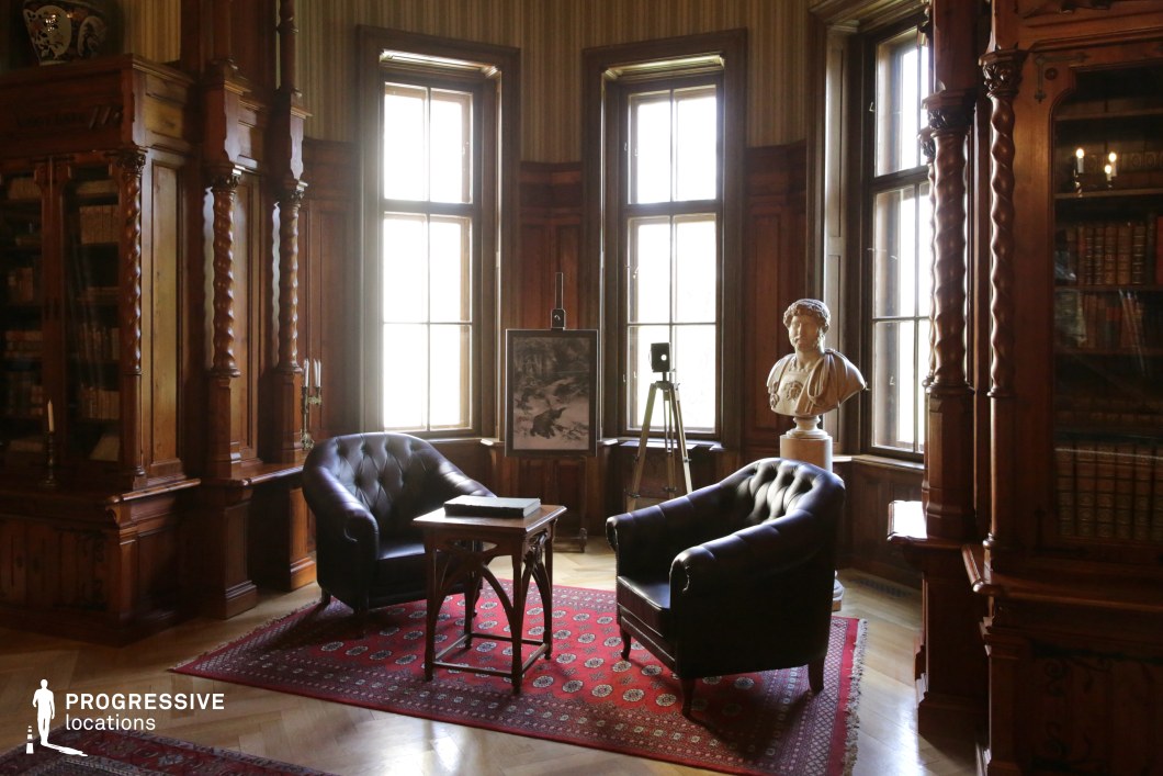 This refined study exudes intellectual sophistication with its dark wood paneling, intricate twisted columns, and built-in bookshelves filled with leather-bound volumes. Large windows framed by wooden moldings allow natural light to softly illuminate the space, highlighting the polished parquet flooring and deep red Persian rug. The room is furnished with two tufted black leather armchairs and a small wooden table, creating an intimate conversational setting. A classical marble bust and an antique-style easel add to the room’s scholarly charm. This location is ideal for period dramas, historical mysteries, or scenes of deep contemplation, offering a timeless and aristocratic atmosphere.