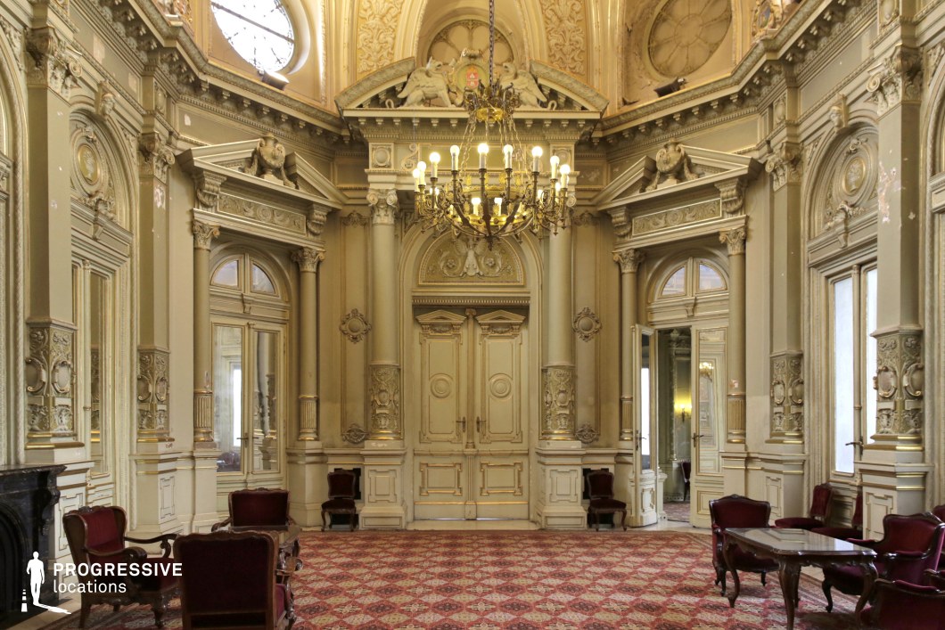 This lavish hall exemplifies neoclassical grandeur, featuring towering columns, intricate moldings, and richly decorated archways. The symmetrical layout is emphasized by large, arched double doors, flanked by ornate pilasters and crowned with sculpted pediments. A grand chandelier hangs from the high, elaborately adorned ceiling, adding to the opulent aesthetic. The muted cream and gold color palette enhances the historical elegance, complemented by antique-style seating and patterned carpeting. This location is ideal for filming period dramas, aristocratic settings, or historical thrillers, offering a regal and timeless ambiance that suits narratives of power, intrigue, and luxury.