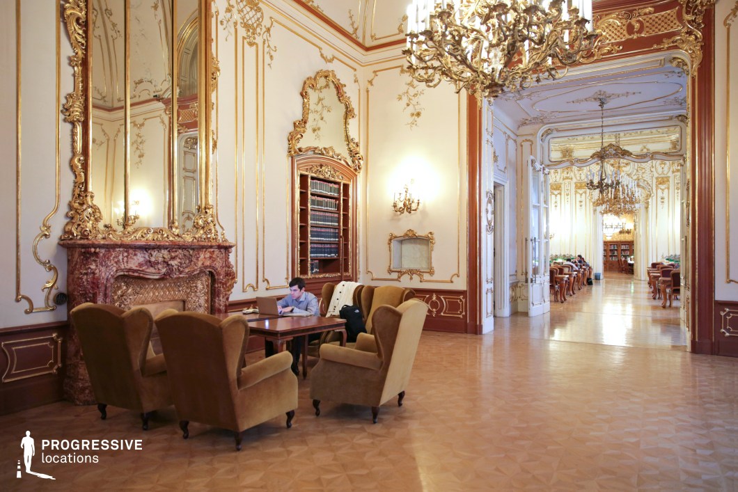 This exquisitely designed Rococo-style library lounge features intricate gold moldings, tall mirrors, and grand chandeliers that enhance the sense of refinement and luxury. The marble fireplace, adorned with elaborate carvings, serves as a central focal point, while built-in bookshelves filled with leather-bound volumes add an intellectual ambiance. Plush velvet armchairs and polished wooden tables create an inviting yet sophisticated atmosphere. The open doorway leads into a secondary reading hall, extending the grandeur of the space with additional chandeliers and decorative wall panels. This location is ideal for filming aristocratic salons, historical debates, or secretive intellectual gatherings, making it a perfect setting for period dramas, classic literary adaptations, or high-society intrigue.