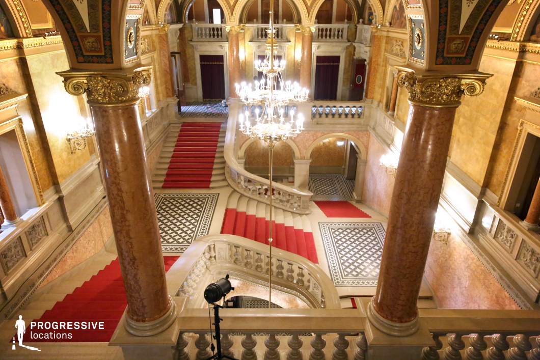 This magnificent neoclassical staircase features sweeping curves, grand marble balustrades, and rich red carpeting that enhances the sense of luxury and formality. Towering marble columns with gilded capitals frame the space, supporting elaborately decorated vaulted ceilings adorned with intricate frescoes and gold detailing. The geometric mosaic flooring contrasts beautifully with the polished stone surfaces, while chandeliers and ornate wall sconces illuminate the space, casting a warm, majestic glow. This location is ideal for filming dramatic arrivals, aristocratic encounters, or high-stakes power struggles in historical dramas, period pieces, or lavish intrigue-filled thrillers.