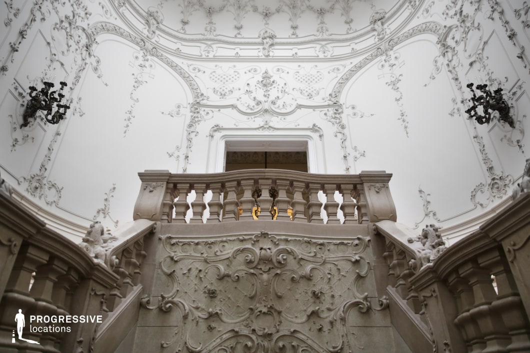 This grand Baroque-style staircase exudes elegance with its elaborate stone balustrades and finely carved reliefs featuring floral and geometric motifs. The surrounding white walls are adorned with intricate stucco decorations, creating a sense of depth and refinement. The symmetry of the architecture is enhanced by the curved railing, leading to a central landing framed by a sculpted archway. Dark, wrought-iron wall sconces contrast against the light-colored background, adding a dramatic touch. This opulent setting is ideal for historical films, period dramas, or fantasy productions, offering a regal and timeless backdrop suitable for scenes of aristocratic life, intrigue, or high-society encounters.