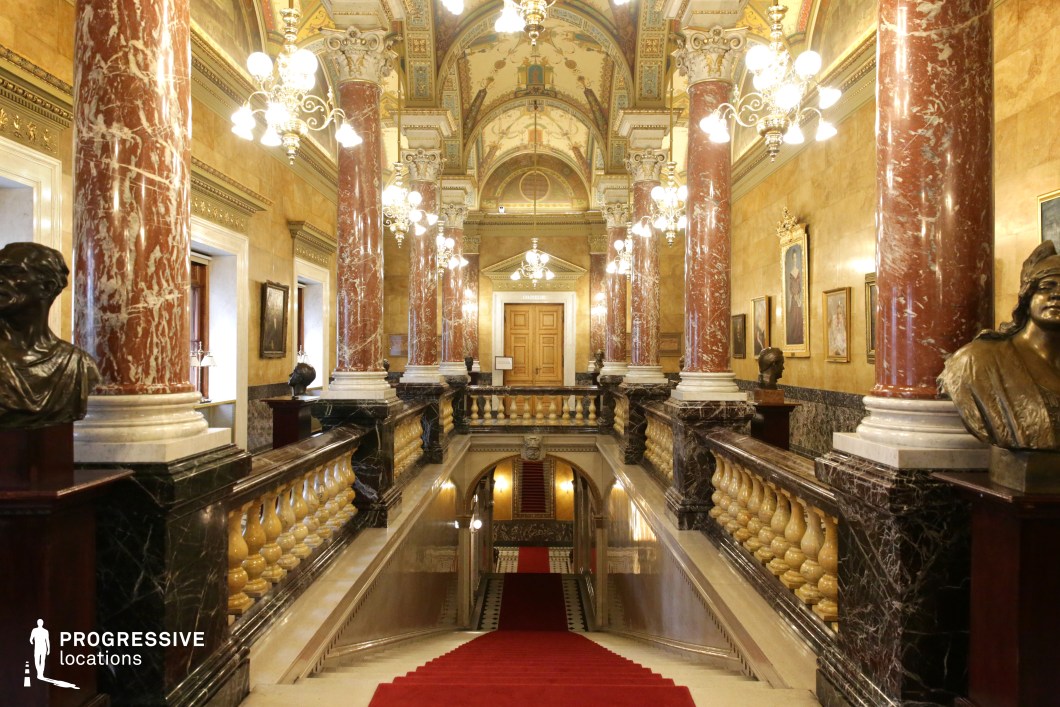This grand neoclassical staircase exudes opulence with its towering red marble columns, ornate gilded ceiling, and intricate wall moldings. The red-carpeted stairs lead upwards towards an imposing set of wooden double doors, framed by decorative arches and illuminated by multiple vintage-style chandeliers. The polished stone balustrades, adorned with golden balusters, complement the rich architectural detailing, while bronze busts and classical paintings add a historical depth to the space. The symmetrical layout and lavish materials create a regal, authoritative atmosphere, making this location ideal for filming dramatic power struggles, aristocratic intrigues, or high-stakes confrontations in historical dramas, political thrillers, or period epics.