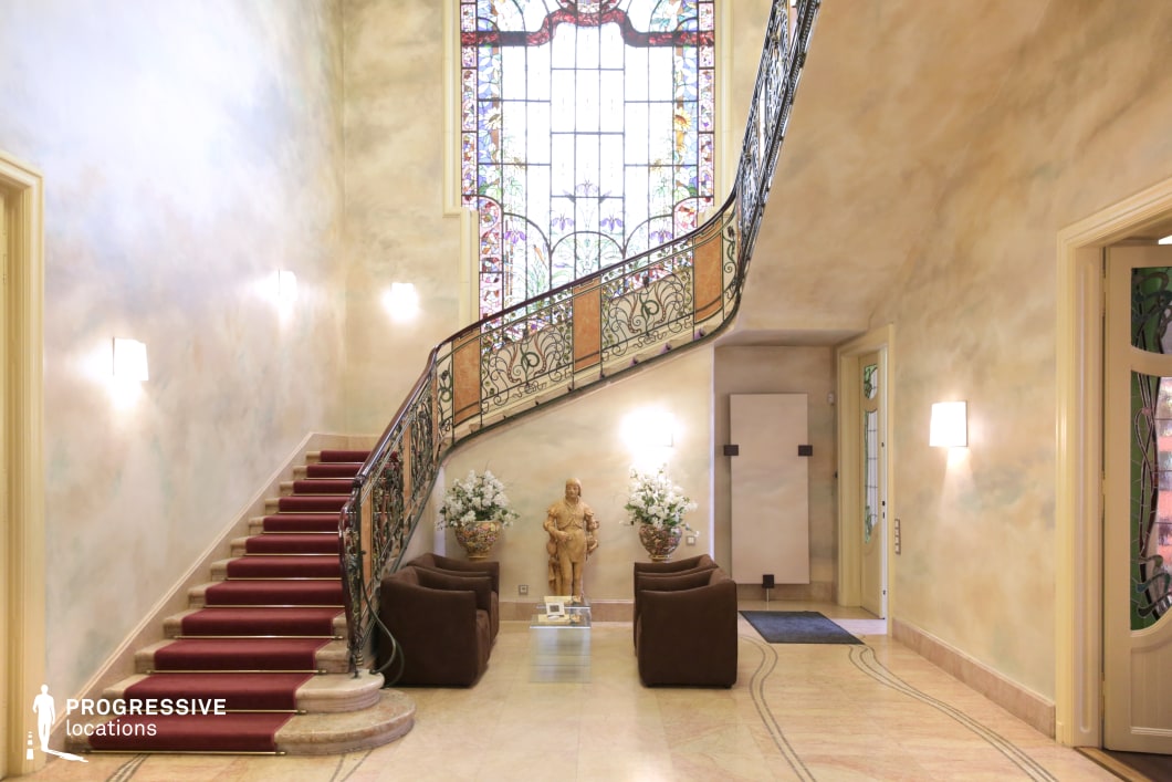 This refined Art Nouveau interior features a gracefully curved staircase with an ornate wrought-iron balustrade adorned with intricate floral motifs. The soft pastel-hued walls and marble flooring create a warm, luminous atmosphere, while a deep red carpet runner adds a touch of classic sophistication. The highlight of the space is a grand stained-glass window, allowing natural light to filter through in a cascade of colors, enhancing the artistic ambiance. Delicate decorative elements, including sculptural accents and floral arrangements, contribute to the room’s harmonious elegance. This location is ideal for filming period dramas, aristocratic settings, or sophisticated intrigue, offering a visually striking and refined environment.