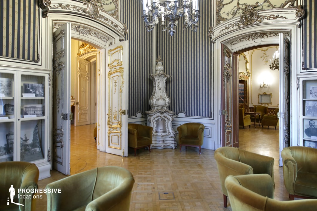 This stunning Rococo-style salon showcases a refined combination of intricate gold moldings, tall arched doorways, and sophisticated striped wallpaper in silver and navy hues. The room is centered around an ornately carved white ceramic fireplace, flanked by mustard velvet armchairs that add a warm contrast to the cool-toned walls. The grand double doors, adorned with gilded carvings, lead into adjoining opulent rooms, enhancing the sense of depth and continuity. Glass display cases filled with historical photographs add a museum-like ambiance, while a classical chandelier casts an elegant glow over the parquet flooring. This location is ideal for filming aristocratic salons, literary discussions, or secretive encounters in historical dramas, period mysteries, and refined thrillers.