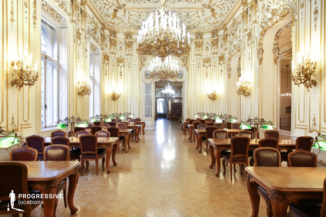 This grand reading hall showcases a stunning Baroque-inspired interior with elaborately carved gold moldings, high arched windows, and an elegant parquet floor. The walls are adorned with intricate gilded ornamentation, complemented by opulent chandeliers and classic wall sconces that illuminate the space with a warm glow. Rows of polished wooden tables with vintage-style chairs create a symmetrical, refined atmosphere, further accentuated by traditional green-shaded library lamps. The combination of luxury, symmetry, and historical grandeur makes this location ideal for filming period dramas, aristocratic settings, historical documentaries, or mystery and suspense films requiring a sophisticated backdrop.