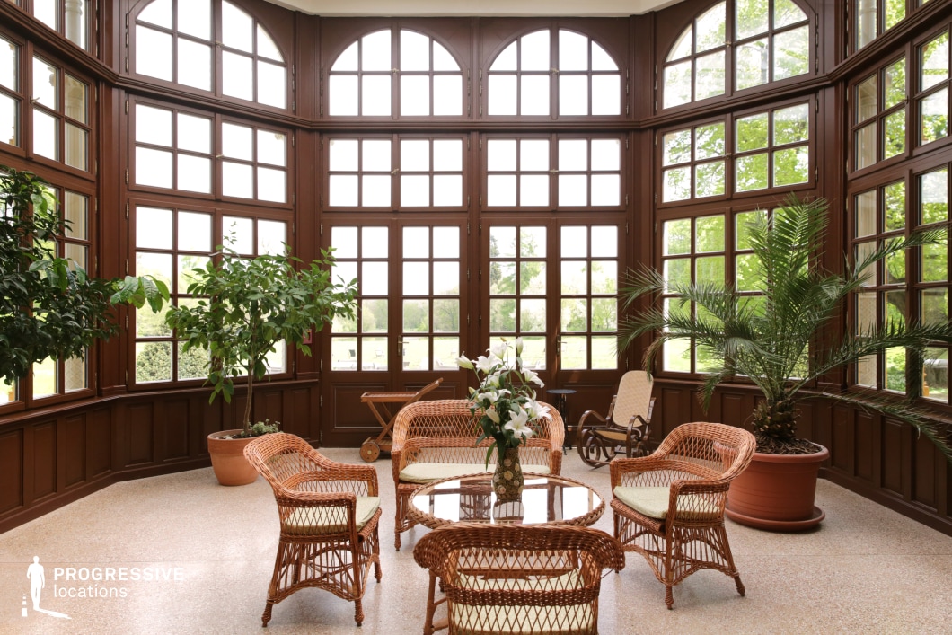 This beautifully designed conservatory features expansive wooden-framed windows that create a bright and airy atmosphere, seamlessly blending the indoors with the surrounding greenery. The arched upper windows add a classic touch, while the dark wood paneling provides a sense of warmth and sophistication. Wicker furniture with soft cushions is arranged around a central glass-top table, enhancing the relaxed yet refined ambiance. Large potted plants contribute to the lush, inviting aesthetic. This location is ideal for filming elegant social gatherings, period dramas, or intimate conversations, offering a tranquil and timeless setting with natural light and a serene garden backdrop.