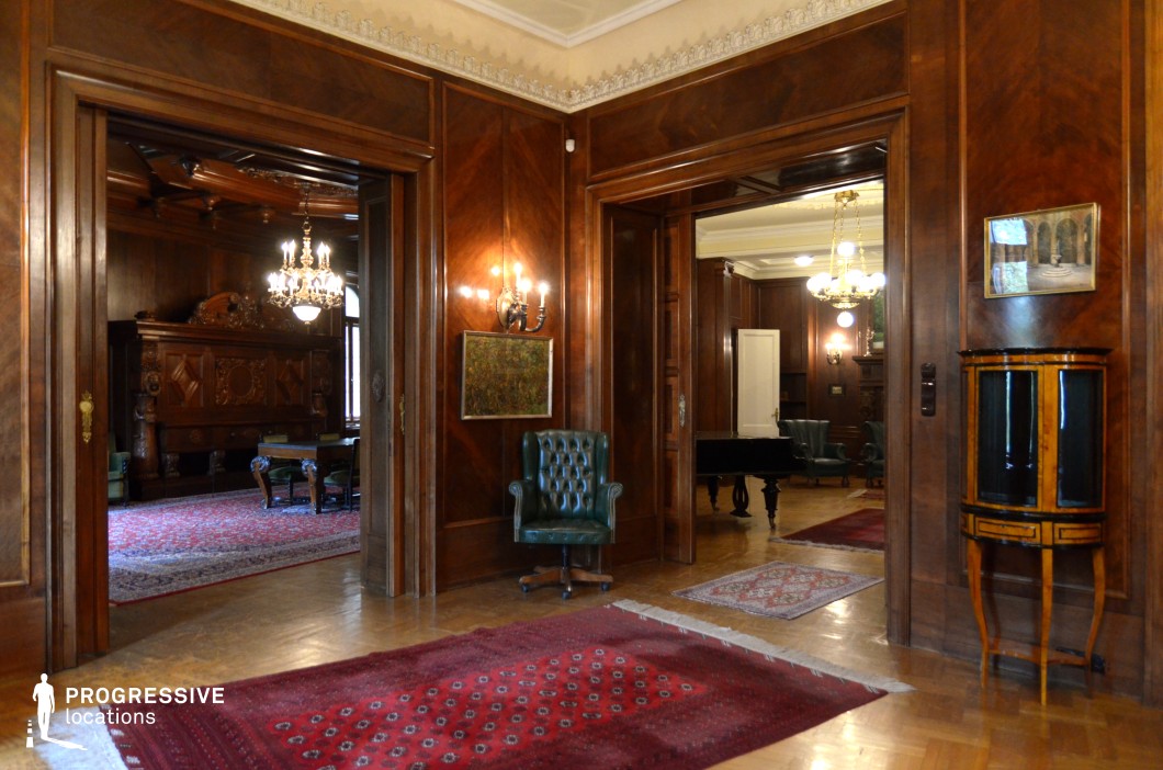 This sophisticated interior features rich, polished wood paneling covering the walls and doorways, creating a refined, classical atmosphere. The space is divided into multiple rooms through large, open doorways, offering a sense of depth and continuity. Ornate chandeliers and wall sconces provide warm illumination, complementing the detailed ceiling moldings. The decor includes luxurious Persian rugs, deep green leather armchairs, and an antique wooden piano, reinforcing the aristocratic charm of the setting. The combination of elegant furnishings and rich textures makes this an excellent location for period dramas, historical films, or mystery thrillers, evoking themes of wealth, power, and intrigue.
