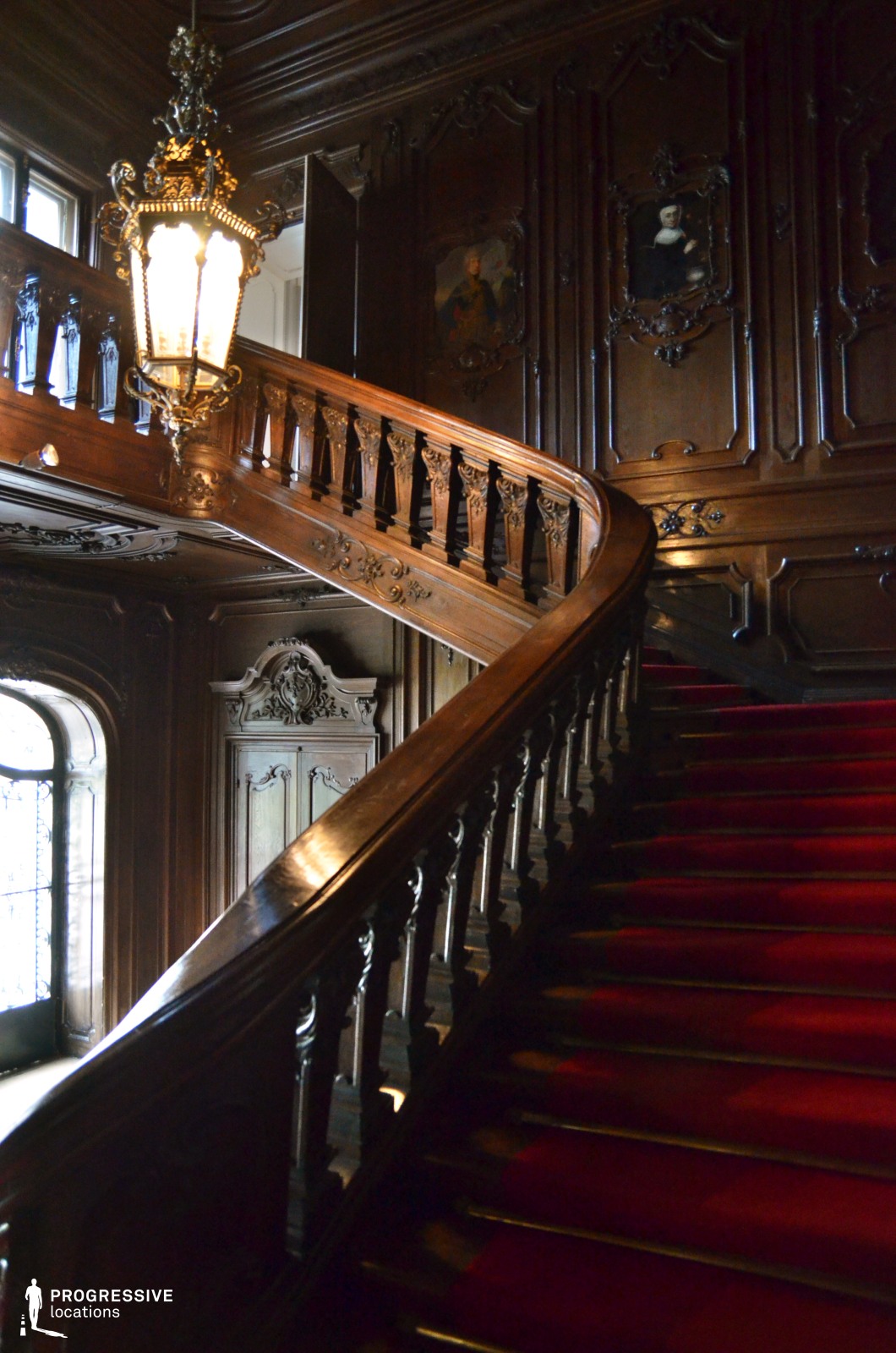 The image showcases a grand, ornately carved wooden staircase with a deep red carpet runner, evoking a sense of opulence and history. The balustrade is adorned with intricate detailing, complementing the dark wood-paneled walls, which feature decorative moldings and embedded portrait paintings in elaborate frames. A large, antique-style lantern hangs from the ceiling, casting a warm glow that enhances the rich textures of the woodwork. The arched window with leaded glass panes allows natural light to filter in, subtly illuminating the space. This setting is ideal for filming period dramas, gothic thrillers, or aristocratic intrigue scenes, providing a visually striking backdrop with a luxurious and timeless ambiance.