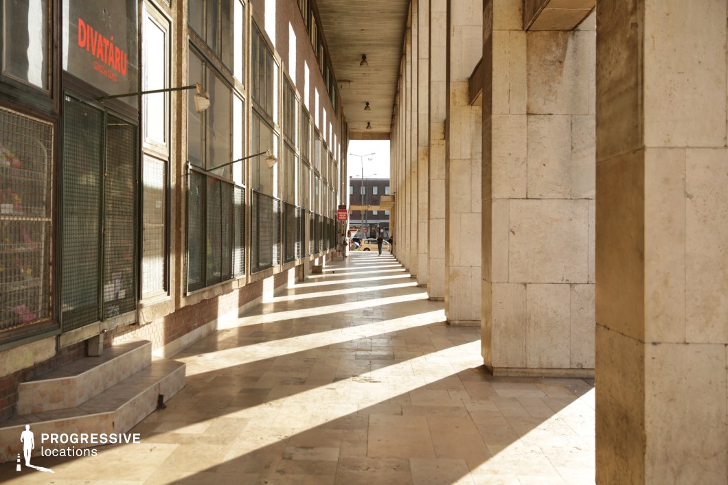 This long, covered arcade is framed by towering stone-clad columns that create a rhythmic pattern of light and shadow across the polished marble floor. The facade features large industrial-style windows, some protected by metal grates, adding to the austere, utilitarian aesthetic. The space exudes a mid-century institutional feel, reminiscent of Soviet-era architecture, with its monumental proportions and structured repetition. The arcade’s perspective leads toward an urban backdrop, adding depth and continuity. This location is ideal for filming historical dramas, political thrillers, or atmospheric urban sequences, making use of its strong geometric elements and striking contrasts.