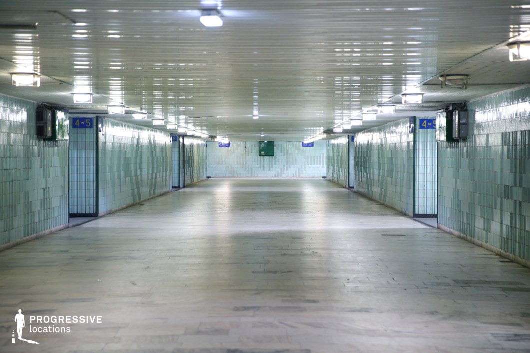 This long, tunnel-like underpass is clad in light blue ceramic tiles, reflecting the artificial overhead lighting that runs symmetrically along the ceiling. The glossy surfaces and pale marble flooring create an almost sterile, institutional atmosphere, while the repetitive signage and doors along the walls suggest a utilitarian transit space. The tunnel’s vanishing point perspective enhances the sense of depth and isolation, making it a visually striking yet slightly unsettling setting. This location is ideal for filming psychological thrillers, dystopian narratives, or suspenseful chase sequences, emphasizing confinement, anonymity, and transition.