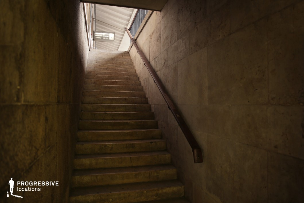 This narrow staircase, enclosed by aged stone walls, leads up to a brighter platform area, creating a strong contrast between light and shadow. The worn stone steps and dark wooden handrail emphasize the utilitarian and slightly decayed aesthetic, adding a sense of history to the space. The enclosing walls enhance the tunnel-like feel, while the overhead signage hints at its function within a transit hub. The setting is ideal for filming suspenseful or introspective scenes, making it suitable for thriller, noir, or mystery genres. Its confined space and moody lighting create a cinematic backdrop for moments of tension, secrecy, or transition between contrasting environments.