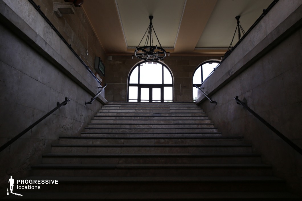 This imposing staircase, framed by aged stone walls and dark metal railings, leads upward toward a set of arched windows that flood the space with natural light. A heavy wrought-iron chandelier hangs from the high ceiling, reinforcing the historical character of the architecture. The worn steps and dimly lit interior contrast with the bright exterior beyond, creating a sense of transition and anticipation. The setting’s symmetry and depth make it an ideal location for filming suspenseful, dramatic, or historical scenes, particularly in thrillers, noir films, or period dramas emphasizing movement between spaces of power, secrecy, or revelation.