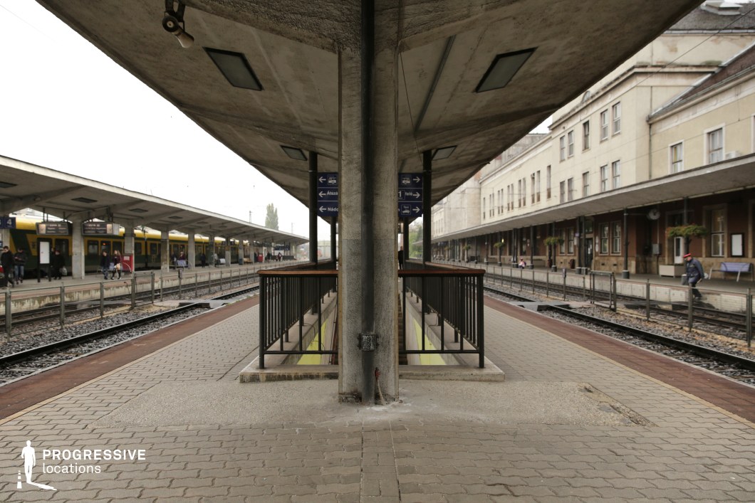 This railway platform features a striking symmetrical composition, dominated by a central concrete pillar supporting a long, overhanging shelter. The brutalist-style canopy, with its raw, weathered concrete surface, extends outward, emphasizing depth and structure. Metal railings and staircases lead to an underground passage, adding layers to the station’s functional design. The surrounding architecture consists of mid-century station buildings with a utilitarian aesthetic, contrasting with the more colorful trains in the background. The combination of repetitive structural elements, linear perspective, and aged materials makes this location ideal for suspenseful dramas, historical films, or dystopian settings that require an industrial or institutional atmosphere.