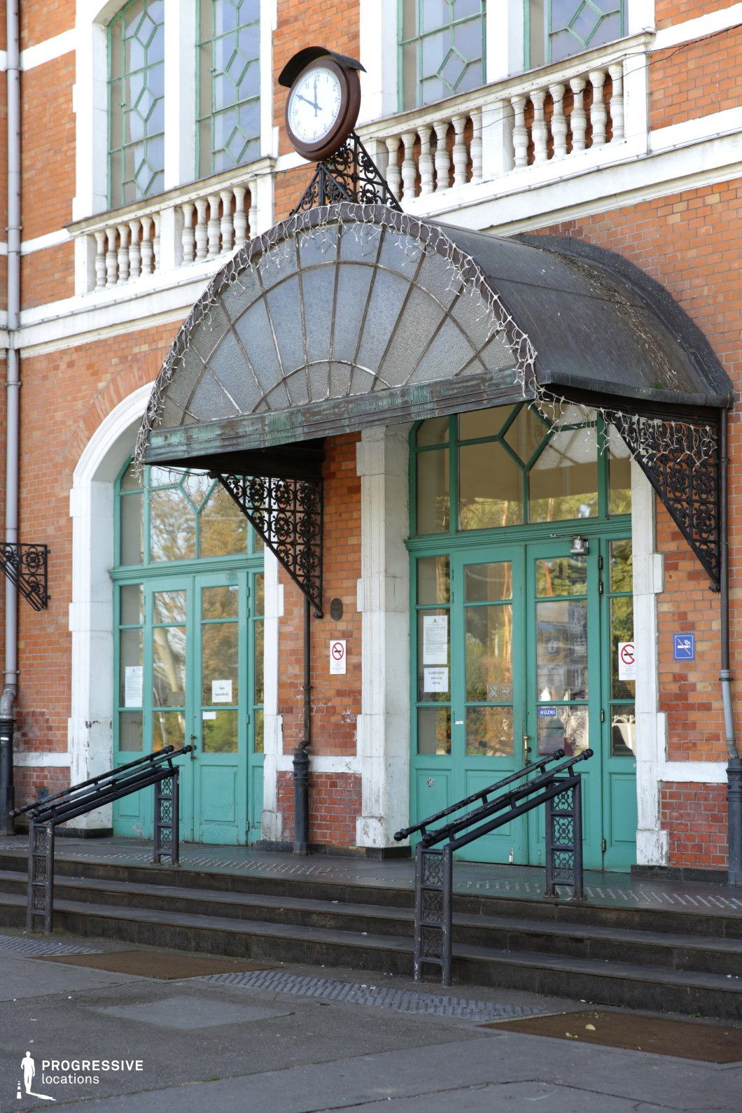 This stunning entrance is a perfect example of Art Nouveau architecture, featuring intricate wrought-iron detailing and beautifully designed doors. The green-painted wooden doors are set against the warm, red brick exterior of the building, with graceful arches and ornamental balconies enhancing its historic charm. Above, a classic clock is mounted in a decorative metal frame, adding a functional yet stylish touch. The gently curved canopy above the stairs is elegantly designed, with a subtle patina of time. The wrought-iron handrails on either side of the steps offer both support and beauty, combining function with aesthetic appeal.