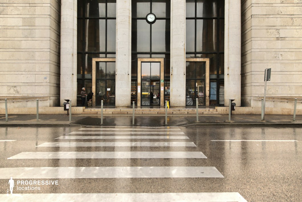 This grand entrance features an imposing neoclassical design, marked by towering stone columns and a large glass facade that reflects the surroundings. The symmetrical layout is emphasized by a central clock above the entrance, adding a formal and institutional feel. The foreground presents a wet pedestrian crossing leading directly to the entrance, enhancing the sense of perspective and directing focus to the building’s authoritative presence. The polished stone exterior, combined with dark metal-framed glass doors, creates a sophisticated yet austere atmosphere. This location is ideal for political dramas, historical narratives, or high-stakes thrillers, evoking themes of power, bureaucracy, and prestige.